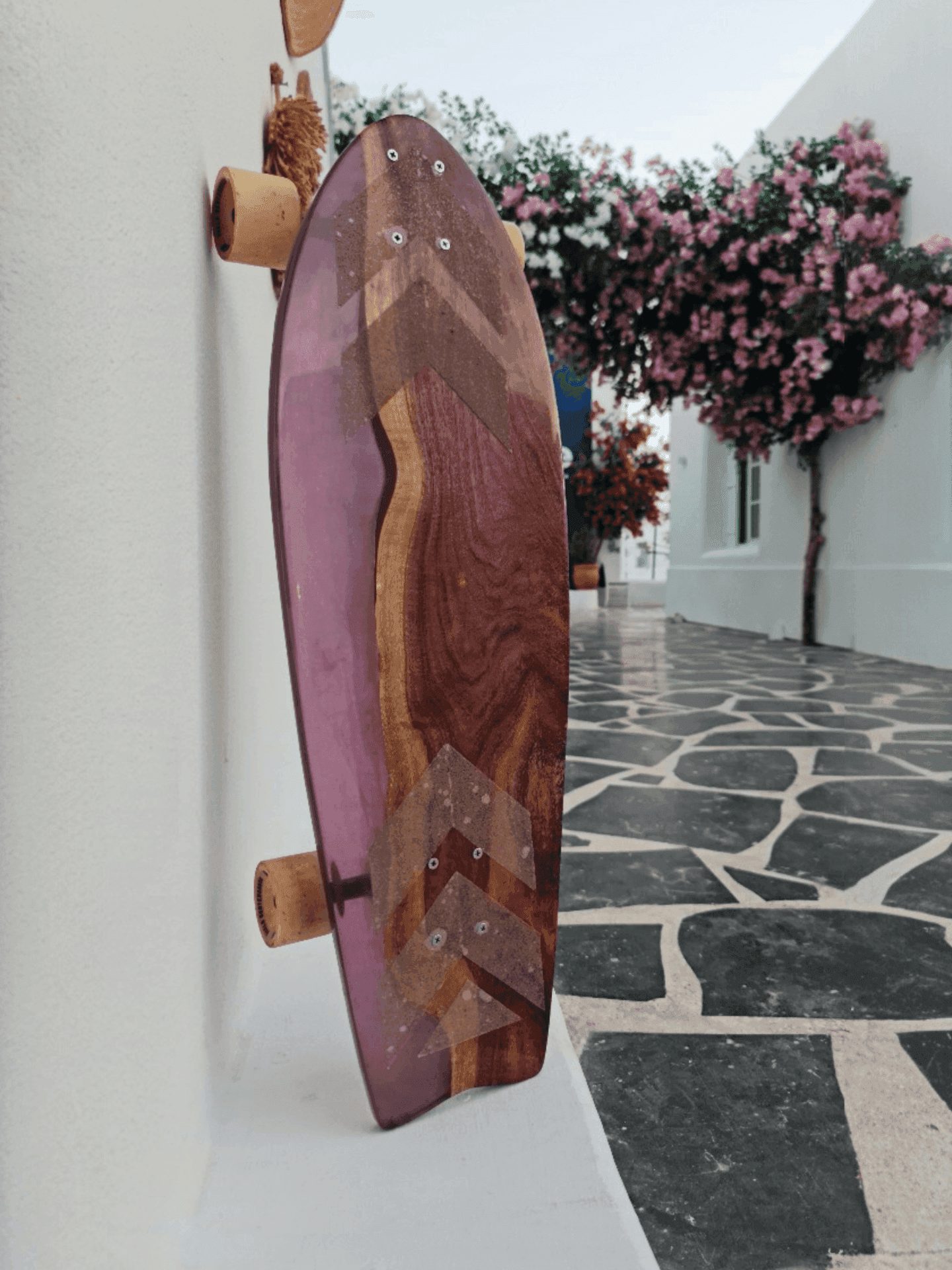 Purple resin chevron longboard leaning on a white wall beside bougainvillea blooms