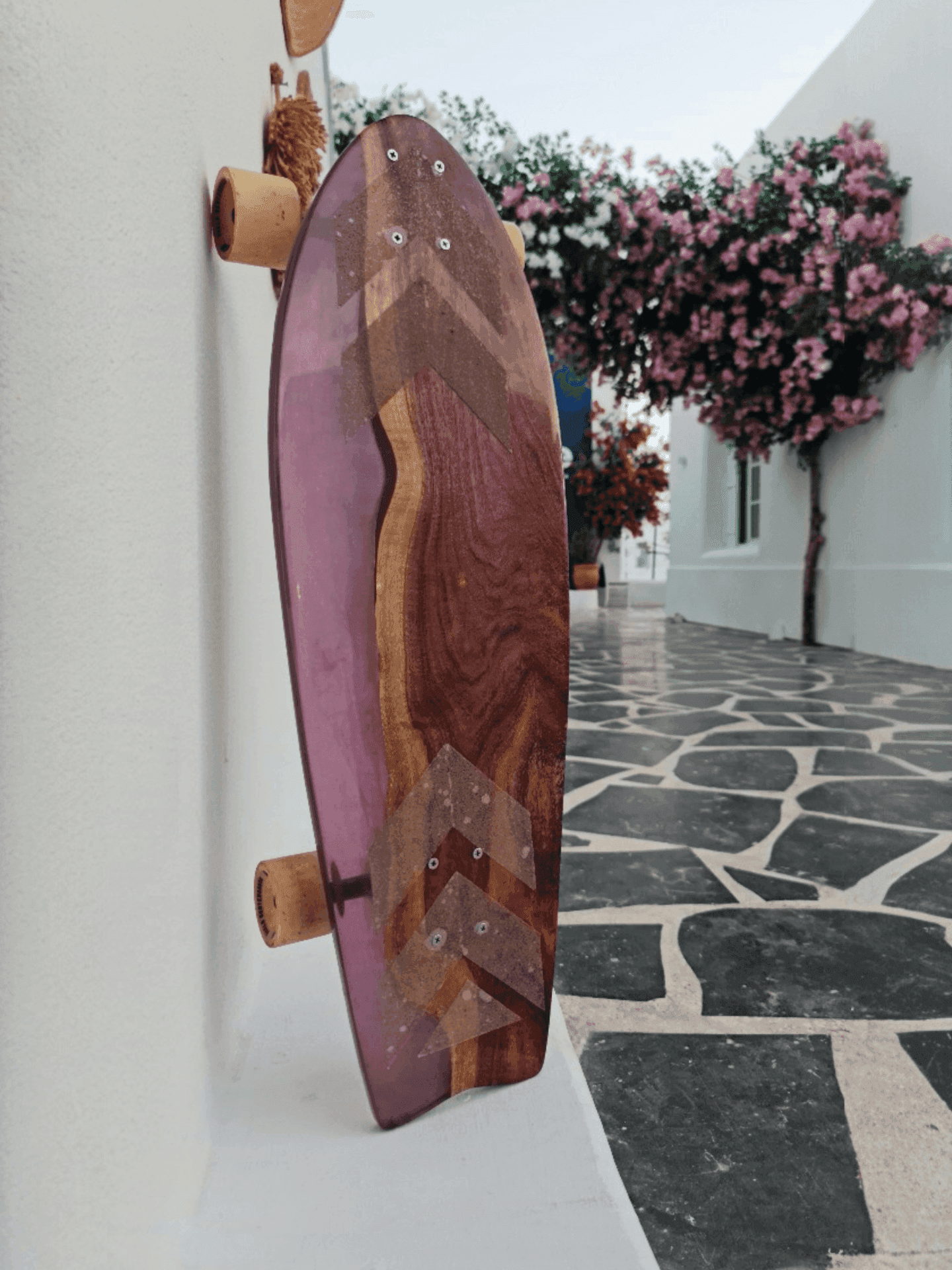 Purple resin chevron longboard leaning on a white wall beside bougainvillea blooms