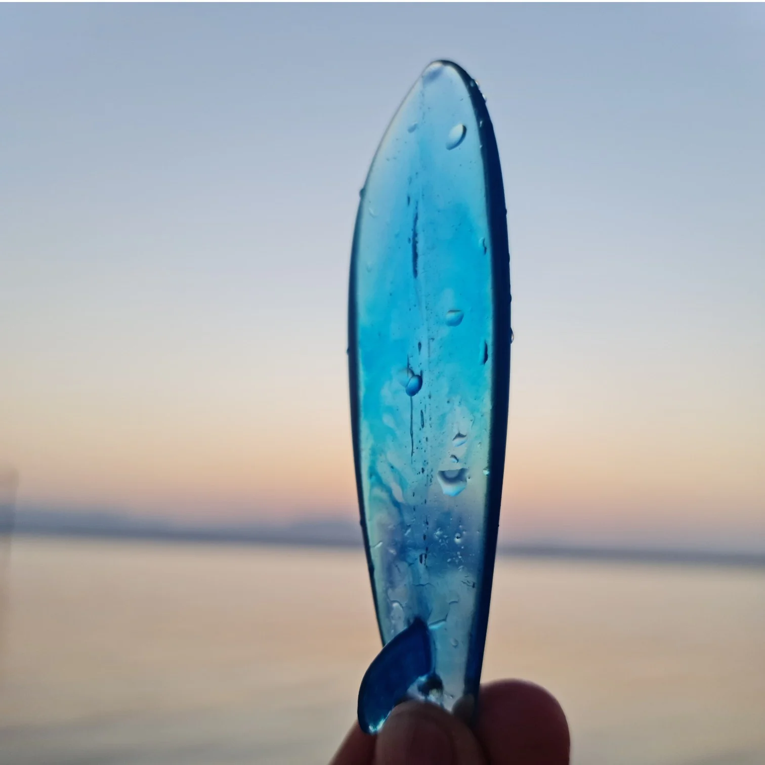 Azure Dawn miniature translucent blue resin surfboard keychain held against an ocean sunrise — view 1